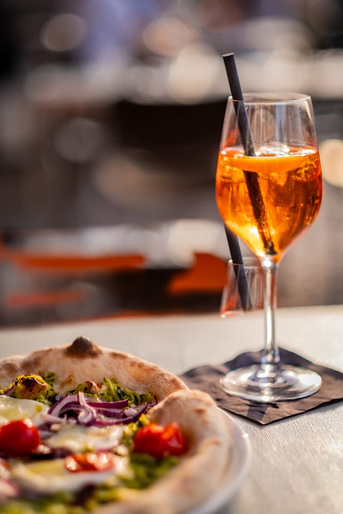 A close-up of an Aperol Spritz cocktail next to a delicious gourmet pizza with blurred background.
